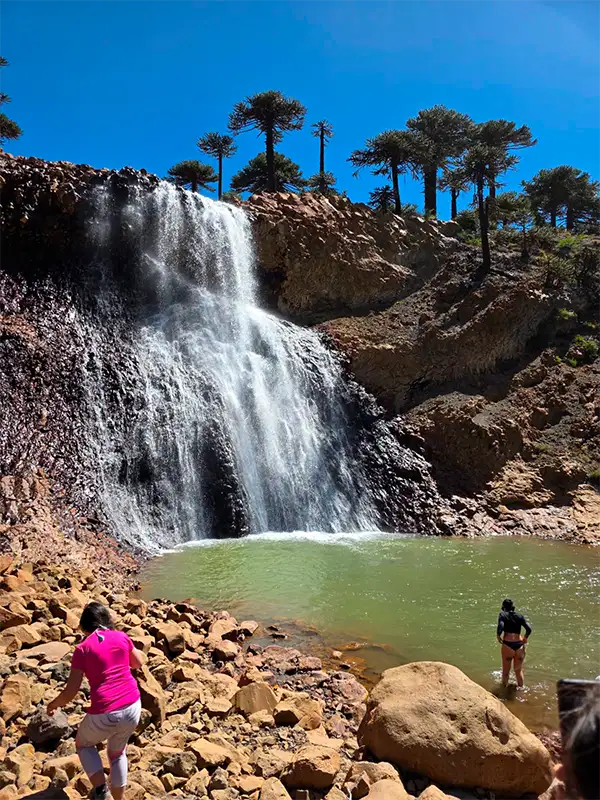 Viajeros refrescándose y entrando al agua en una pileta natural color esmeralda al pie de una cascada rodeada de bosques de araucarias en Caviahue.