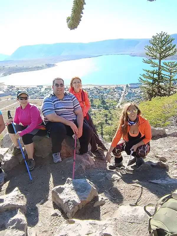 Grupo de cuatro personas haciendo trekking con mochilas y bastones en un mirador con vista al lago y al pueblo de Caviahue. Experiencia de viaje con Vivak Aventuras.