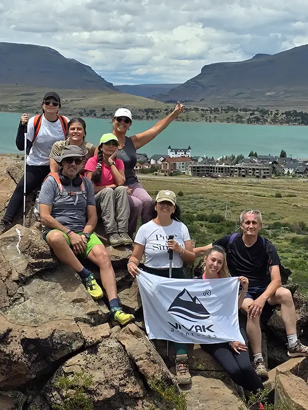 Grupo de excursionistas posando sonrientes con la bandera de Vivak Aventuras en un mirador rocoso con vistas panorámicas al Lago Caviahue y la villa turística de fondo.