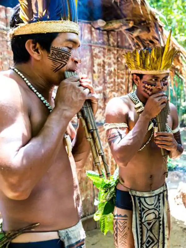 Dos hombres de una comunidad indígena del Amazonas con pinturas faciales tradicionales tocando flautas de pan de madera.