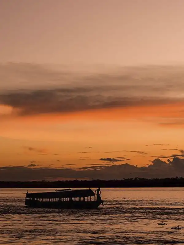 Silueta de un bote tradicional navegando por el inmenso río Amazonas bajo un cielo atardecer naranja.