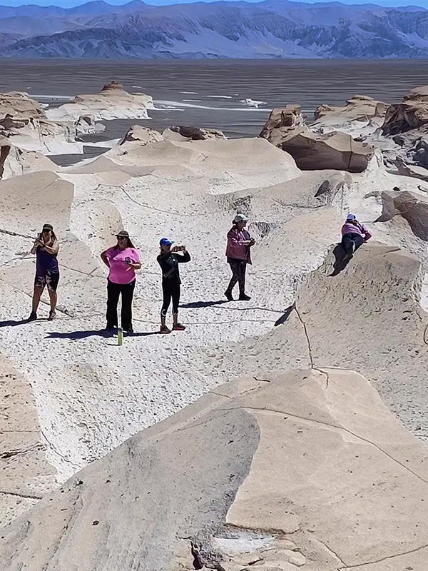 Un grupo de cinco personas vestidas con ropa de trekking colorida explora una formación rocosa de color beige y blanco en el Campo de Piedra Pómez. Una persona se toma una selfie, otra posa en rosa, y otra saca una foto con su celular, todo bajo un sol radiante con montañas al fondo.