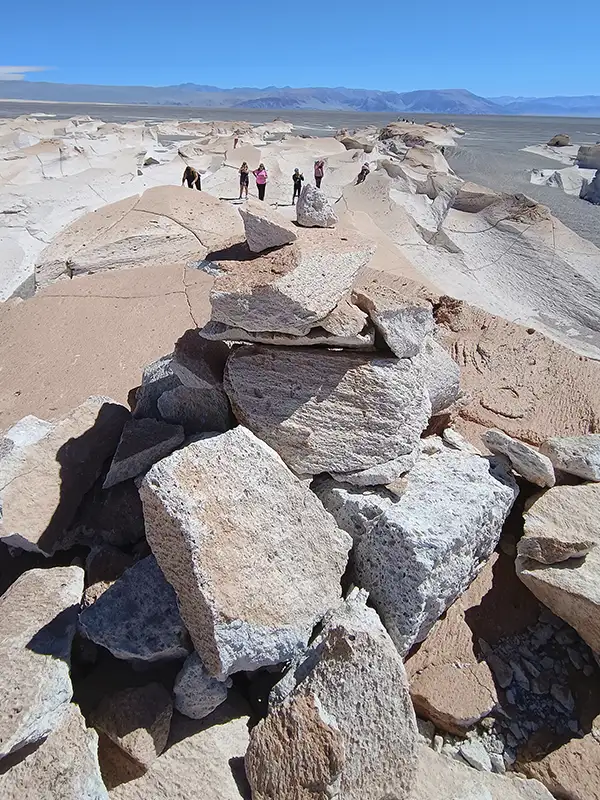 Primer plano nítido de un montículo de rocas de piedra pómez de color beige y blanco, con una textura rugosa y porosa muy definida. Al fondo, un paisaje desértico vasto con pequeñas figuras de personas a lo lejos, bajo un cielo azul sin nubes.
