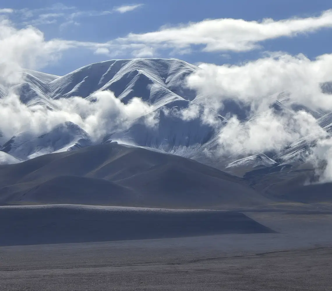 Paisaje de las montañas de la Puna de Catamarca con cimas nevadas y laderas oscuras, parcialmente cubiertas por nubes flotantes, sobre un vasto desierto en primer plano, perfecto como fondo de héroe para un sitio de viajes.