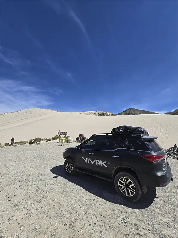 Paisaje panorámico de la Puna en Catamarca mostrando un convoy de camionetas 4x4 transitando por un camino de tierra entre rocas volcánicas, con inmensas montañas y mesas al fondo bajo un cielo nublado.
