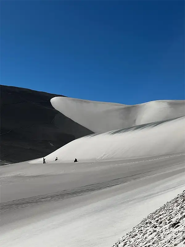 Inmensa duna de arena blanca con forma de cresta en Dunas Blancas, Catamarca, contrastando con cerros oscuros y pequeñas siluetas de personas que muestran su gran escala.