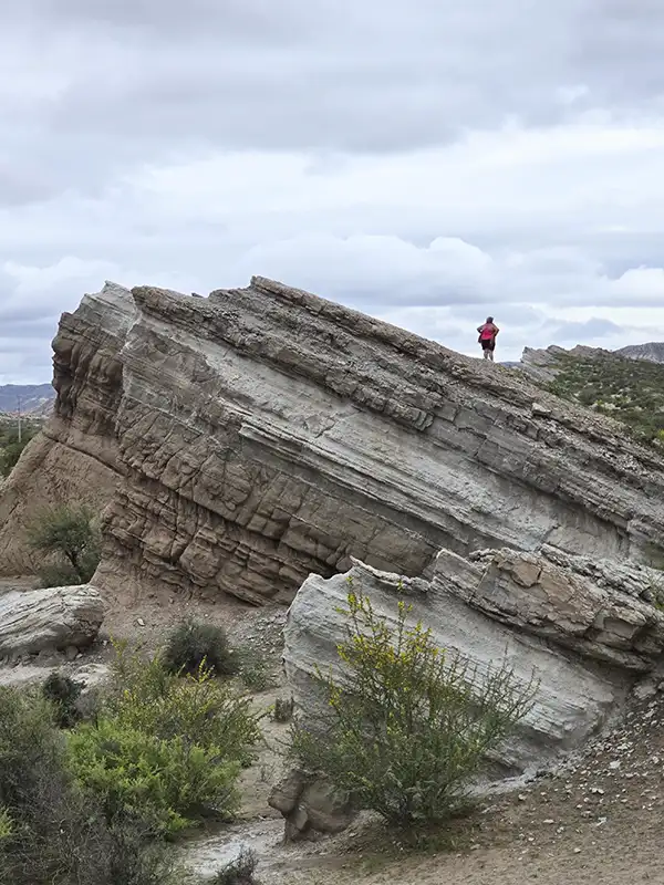 Persona de pie sobre una gran formación rocosa con capas geológicas visibles en el Cañón de Mirihuaca, Catamarca, bajo un cielo nublado.