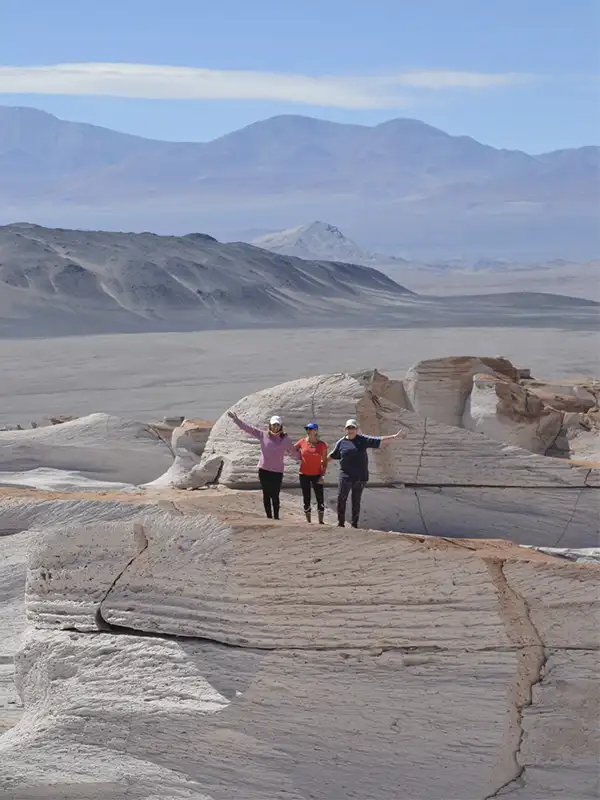 Tres personas celebrando sobre las formaciones de piedra pómez blanca con una vista panorámica del desierto y montañas de Catamarca al fondo.