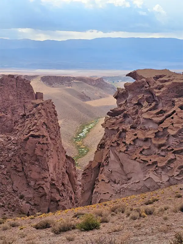 Vista desde lo alto del Cañón de Mirihuaca en Catamarca, donde dos grandes formaciones de roca rojiza enmarcan un valle fértil con un pequeño río al fondo.