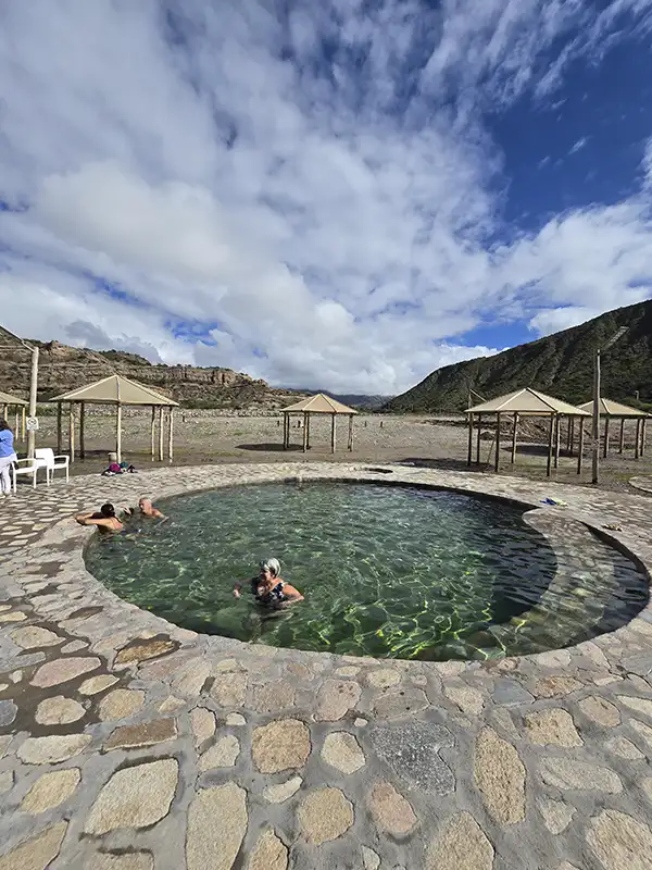 Pileta termal circular de piedra en las Termas de Villa Vil, con personas disfrutando del agua y un paisaje de montañas y nubes de fondo.