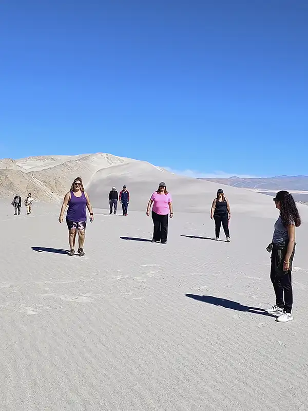 Turistas caminando sobre dunas de arena blanca en la Puna catamarqueña, con montañas a lo lejos y un horizonte despejado.