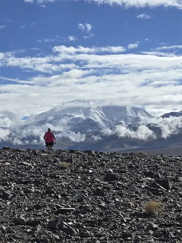 Viajero caminando por un suelo de basalto oscuro frente a un imponente volcán con la cima nevada y rodeado de nubes en la Puna argentina.