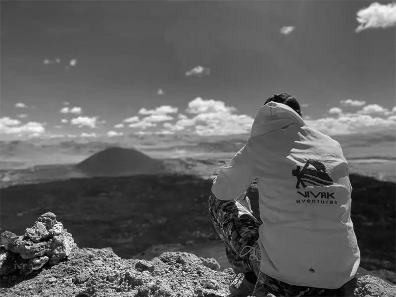 otografía desde la cima de un cerro donde una persona de espaldas, con una campera amarilla fluorescente de "Vivak Aventuras", observa un imponente cono volcánico rodeado de campos de lava negra. El cielo es de un azul intenso con pequeñas nubes blancas, destacando la inmensidad del paisaje de la Puna.