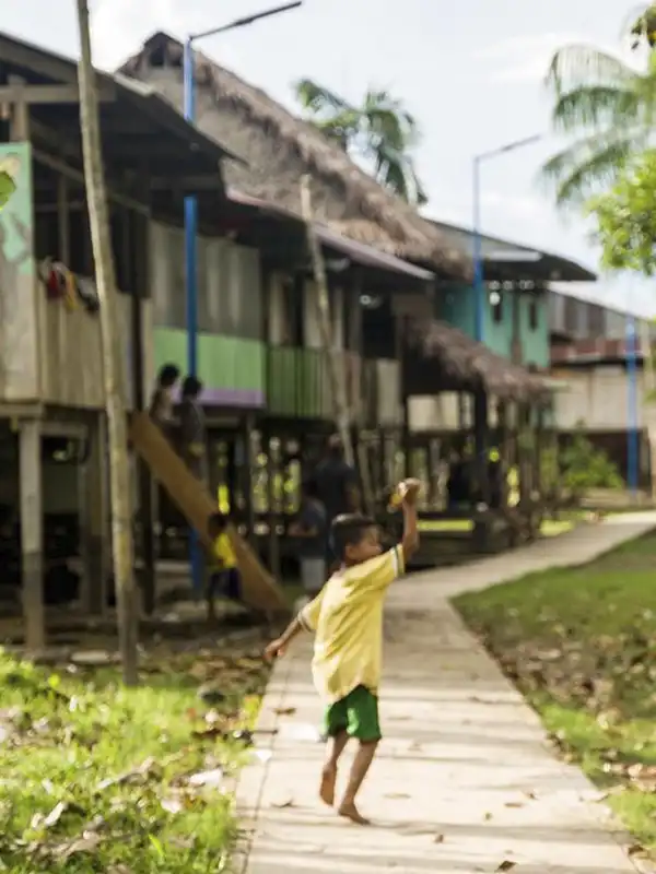 Un niño pequeño y descalzo corre sonriendo por un sendero pavimentado en una comunidad indígena tradicional de la selva amazónica, con palafitos de madera a los lados.