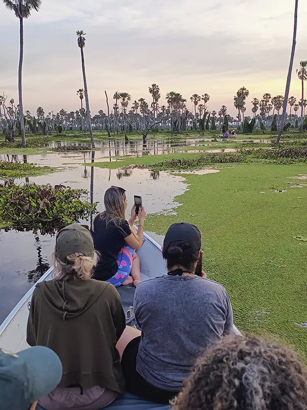 Viajeras fotografiando el paisaje de palmeras y champales reflejados en el agua desde una canoa al atardecer en el Bañado La Estrella, Formosa.