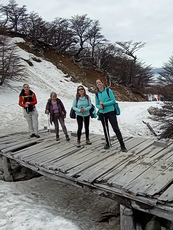 Grupo de viajeros caminando con bastones de trekking sobre un puente de madera rodeado de nieve y bosque de lengas en Ushuaia, Tierra del Fuego.