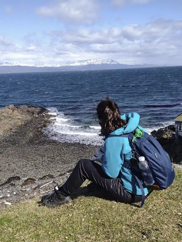 Viajera sentada sobre la costa rocosa contemplando el Canal de Beagle con montañas nevadas al fondo en Ushuaia, Tierra del Fuego.