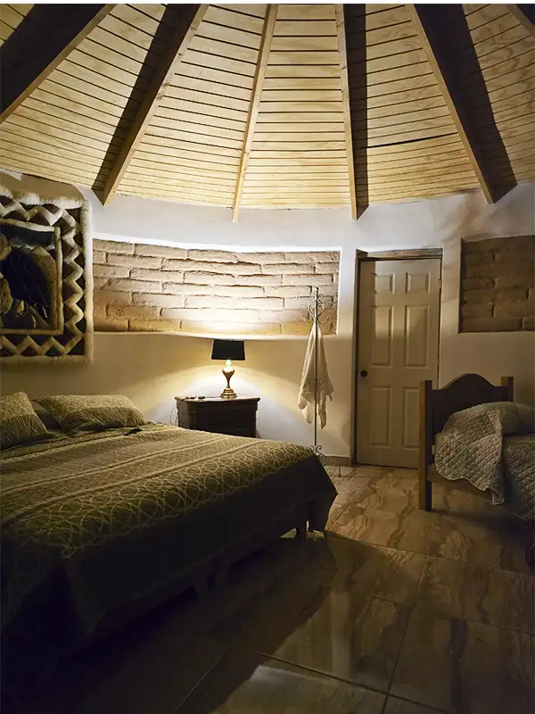 Interior de habitación del hospedaje en Atacama con techo cónico de madera, paredes de piedra y cama doble con iluminación cálida