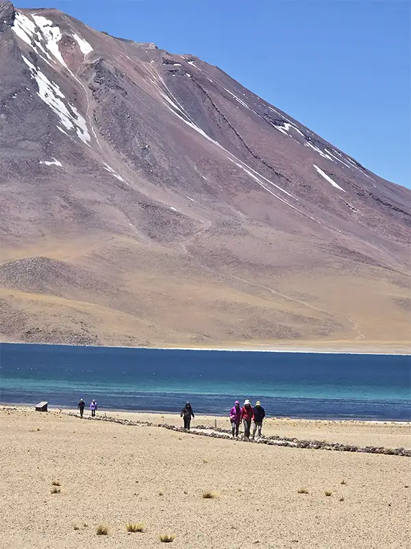 Viajeros caminando a orillas de una laguna altiplánica de agua turquesa con volcán nevado de fondo
