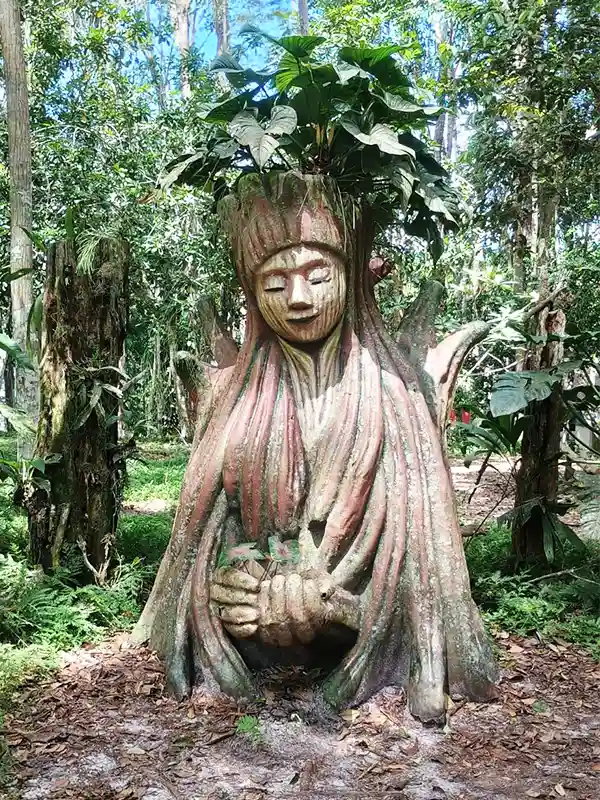 Escultura tallada en un árbol con forma de figura ancestral y planta en la cabeza, rodeada de selva amazónica, Perú.